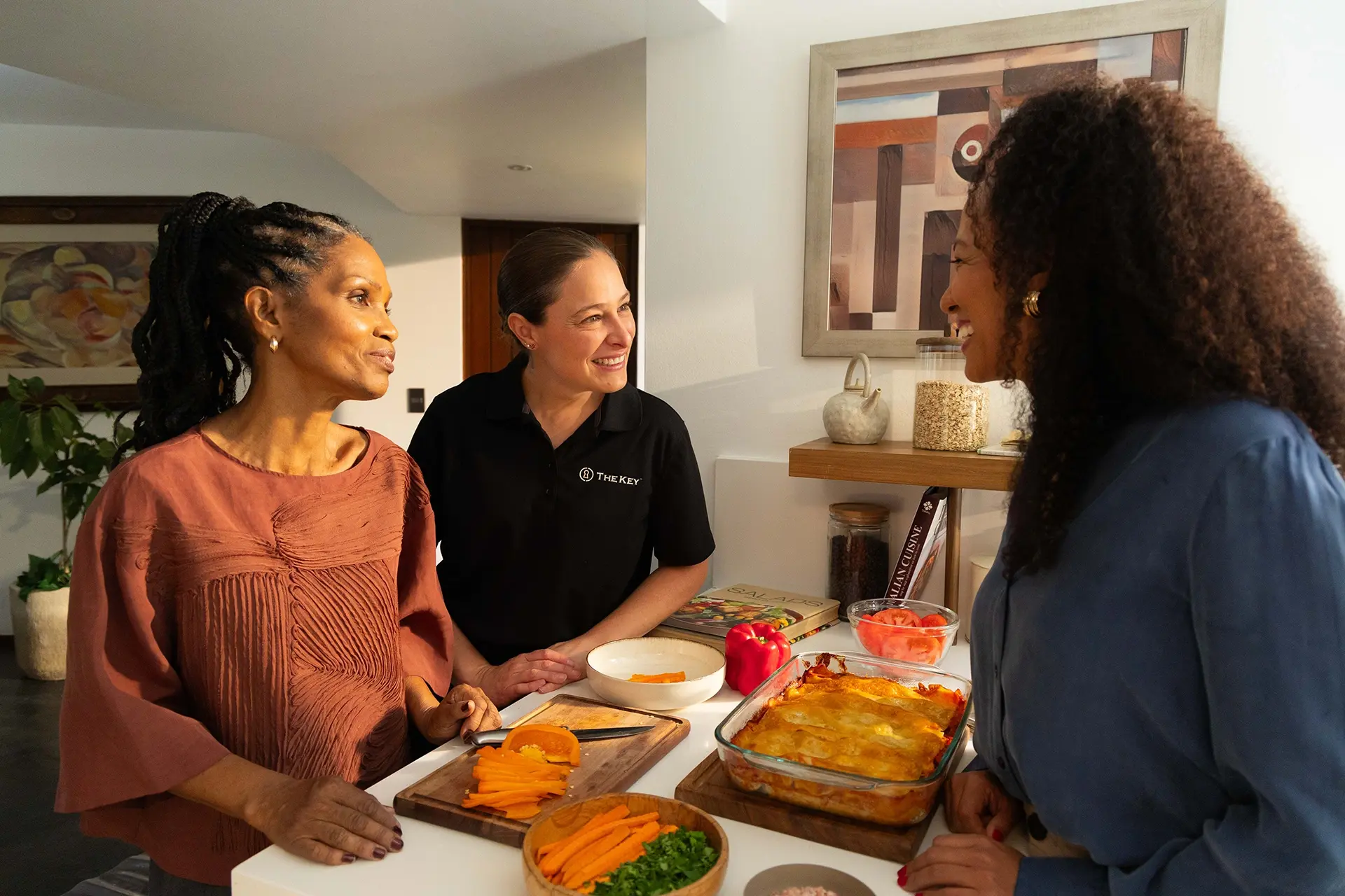 TheKey caregiver conversing warmly with a senior woman and her family member in the kitchen, reflecting the collaborative family partnership and personalized in-home care coordination that TheKey provides